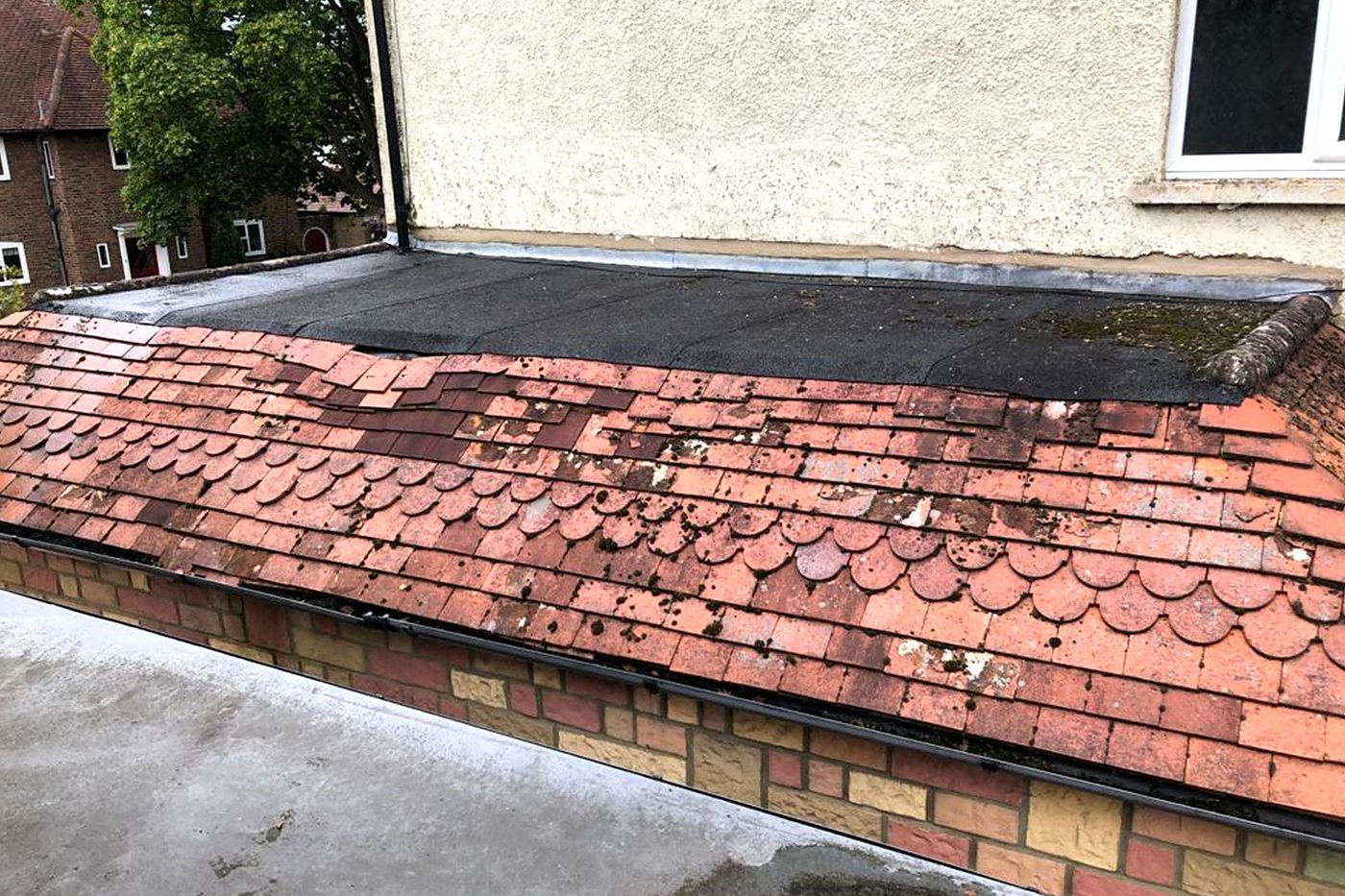 Old plain clay tiles with moss beside a tired felt flat roof